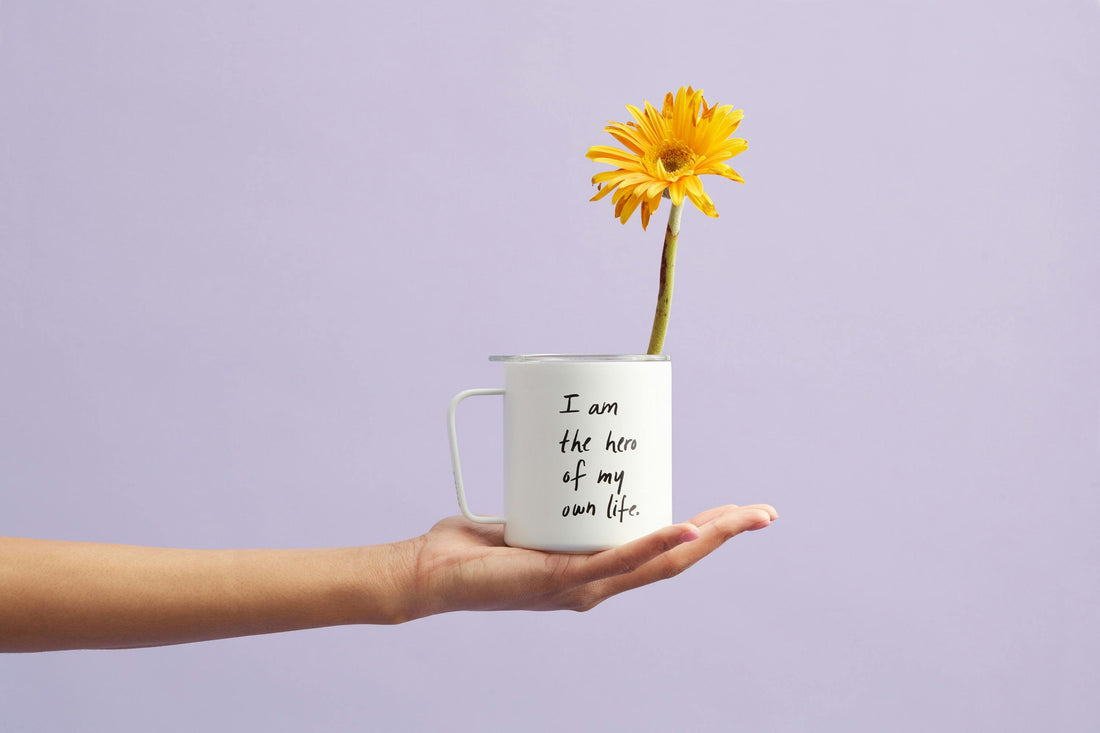 I am the hero of my own life- HaEine gelbe Blume, die aus einer weißen Tasse mit der Aufschrift 'I am the hero of my own life' wächst. Die Tasse wird von einer ausgestreckten Hand vor einem zarten lila Hintergrund gehalten.